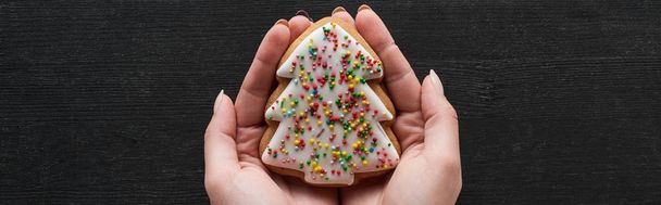 cropped view of woman holding Christmas tree cookie in hands on black wooden table, panoramic shot - Foto, Bild