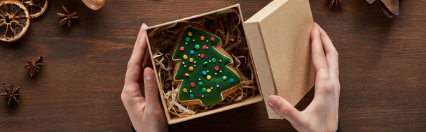 oříznutý pohled na otevírací box ženy s vánoční stromečkem sušenkou u dřevěného stolu, panoramatický záběr - Fotografie, Obrázek