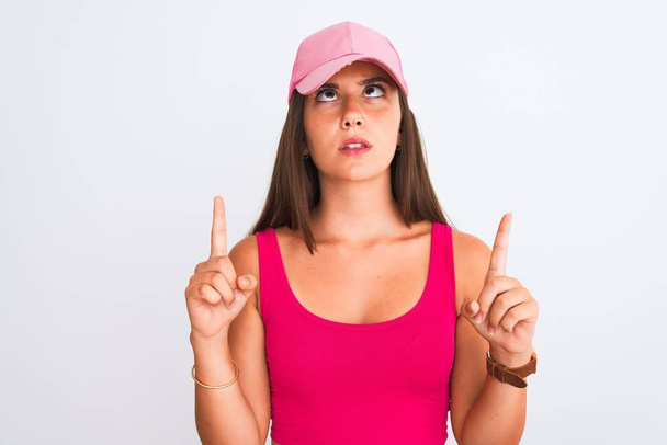 Jeune belle fille portant rose casual t-shirt et casquette sur fond blanc isolé étonné et surpris de regarder vers le haut et pointant avec les doigts et les bras levés
. - Photo, image
