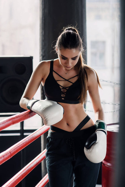 Tired sports girl leaned on red ropes on boxing ring, and have a rest after hard training in black loft gym. Healthy and sporty lifestyle concept. - Foto, afbeelding