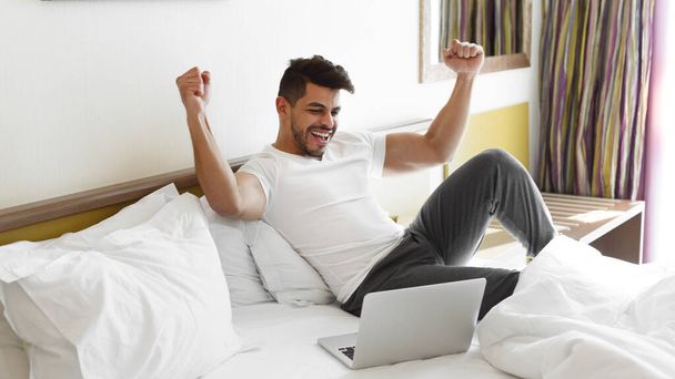 Jovem comemorando o sucesso, sentado na cama, usando laptop
 - Foto, Imagem