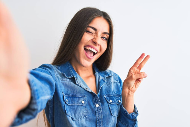 Beautiful woman wearing denim shirt make selfie by camera over isolated white background smiling with happy face winking at the camera doing victory sign with fingers. Number two. - Foto, Bild