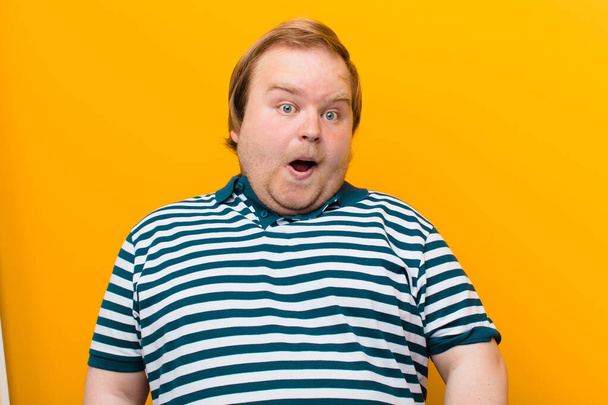 young big size man feeling terrified and shocked, with mouth wide open in surprise against orange wall - Fotoğraf, Görsel