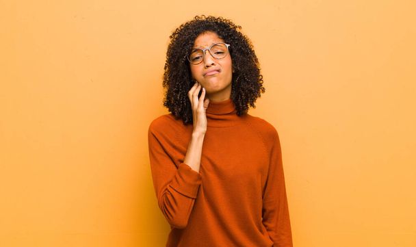 young pretty black woman holding cheek and suffering painful toothache, feeling ill, miserable and unhappy, looking for a dentist against orange wall - Фото, изображение