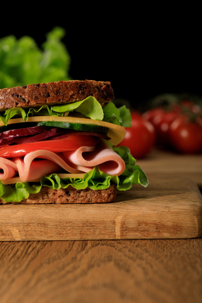 close up view of fresh sandwich with lettuce, ham, cheese, bacon and tomato on wooden cutting board isolated on black - Foto, immagini