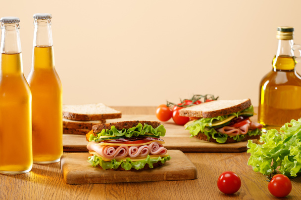 selective focus of fresh sandwiches with lettuce, ham, cheese, bacon and tomato near bottles of beer at wooden table isolated on beige - Photo, image