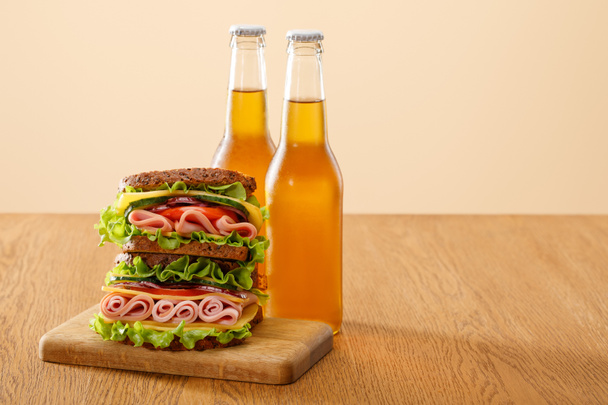 fresh sandwich with lettuce, ham, cheese, bacon and tomato near bottles of beer at wooden table isolated on beige - Photo, image