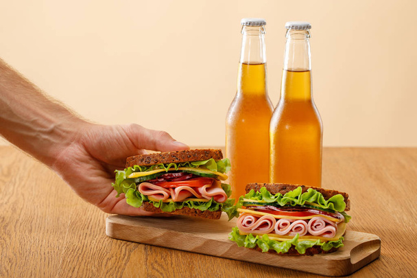 cropped view of man holding fresh sandwich with lettuce, ham, cheese, bacon and tomato near bottles of beer at wooden table isolated on beige - Fotografie, Obrázek