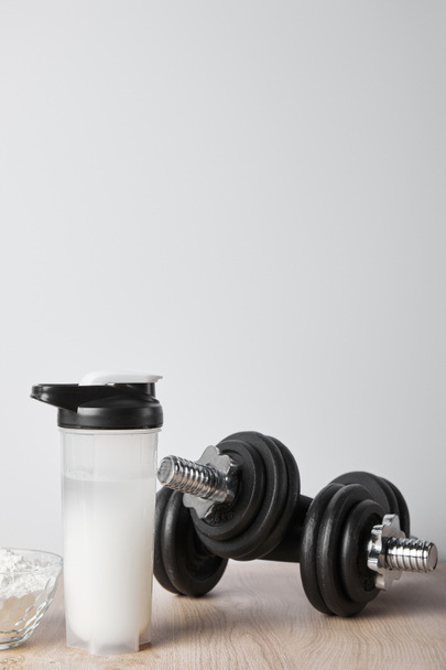 спортивная бутылка с протеиновым коктейлем рядом с гантелями, изолированными на сером
 - Фото, изображение