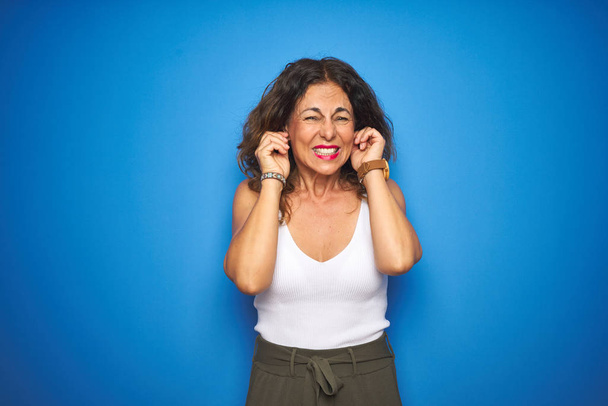 Middle age senior woman with curly hair standing over blue isolated background covering ears with fingers with annoyed expression for the noise of loud music. Deaf concept. - Photo, Image