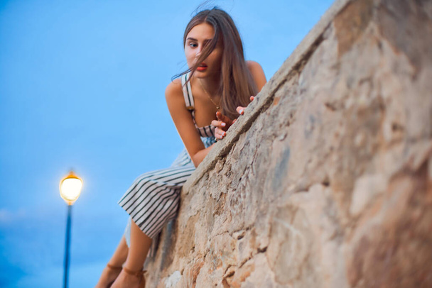 Beautiful woman wearing striped overalls and stylish sandals sitting on a stone wall . - Photo, Image
