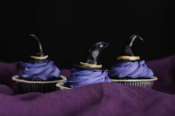 tasty Halloween cupcakes with blue cream and decorative witch hats on purple cloth isolated on black - Foto, Bild