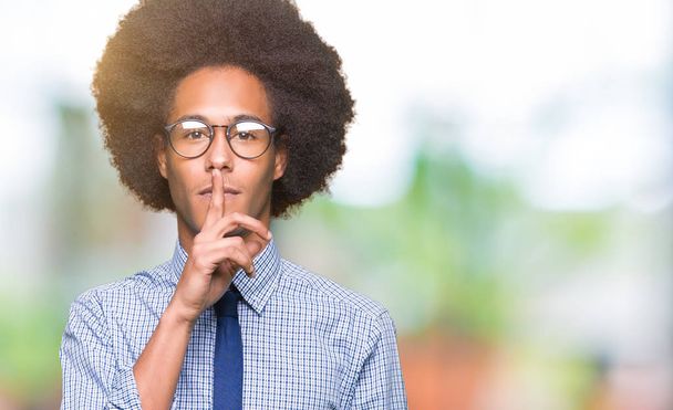 Молодой африканский бизнесмен с афроволосами в очках просит тишины с пальцем на губах. Молчание и тайная концепция
. - Фото, изображение