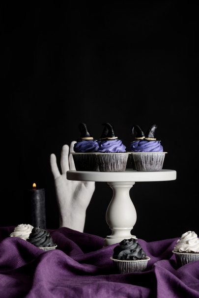 tasty Halloween cupcakes on white stand near burning candles and decorative hand on purple cloth isolated on black - Fotoğraf, Görsel
