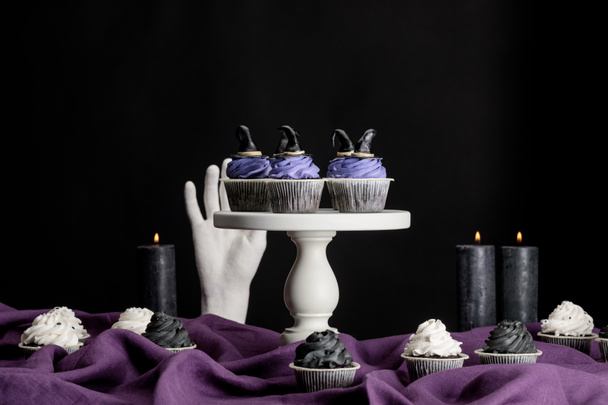 chutné Halloween cupcakes na bílém stojanu v blízkosti hořící svíčky a dekorativní ruku na fialové látky izolované na černé - Fotografie, Obrázek