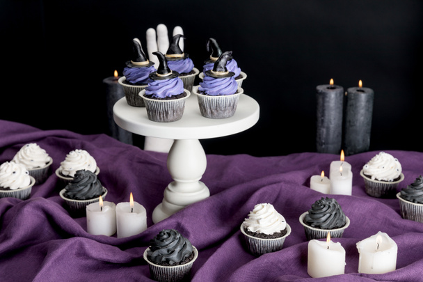 tasty Halloween cupcakes on white stand near burning candles on purple cloth isolated on black - Fotó, kép