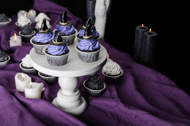 tasty Halloween cupcakes on white stand near burning candles on purple cloth isolated on black - Zdjęcie, obraz