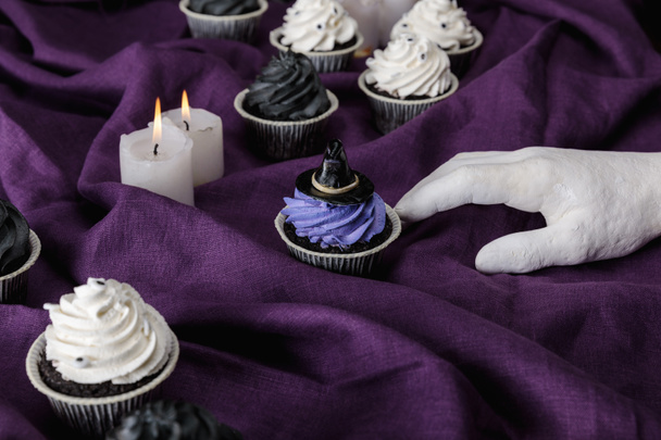 decorative hand near tasty Halloween cupcakes and burning candles on purple cloth - Photo, Image