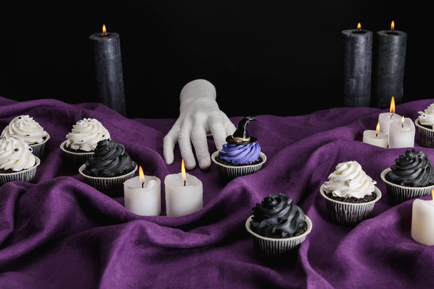 decorative hand near tasty Halloween cupcakes and burning candles on purple cloth isolated on black - Фото, изображение
