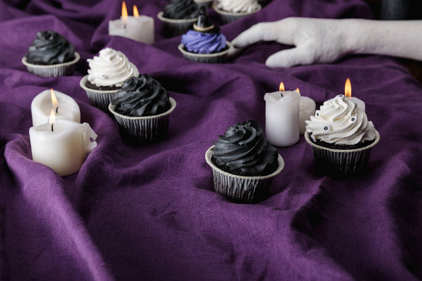 decorative hand near tasty Halloween cupcakes near burning candles on purple cloth - Fotoğraf, Görsel
