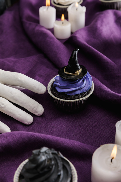 декоративна рука біля смачного кексу на Хеллоуїн з блакитним кремом та декоративним капелюхом відьми біля палаючих свічок на фіолетовій тканині
 - Фото, зображення