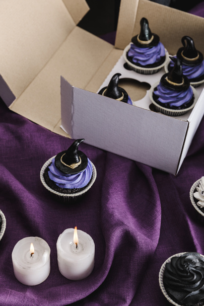 tasty Halloween cupcakes with blue cream and decorative witch hats in box near burning candles on purple cloth - Photo, Image