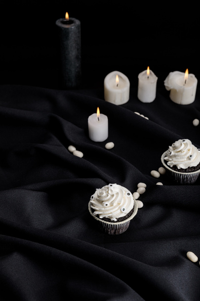 tasty Halloween cupcakes with white cream near burning candles on black cloth - Foto, afbeelding