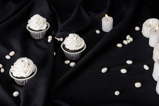 smakelijke Halloween cupcakes met witte room in de buurt van brandende kaarsen op zwarte doek - Foto, afbeelding