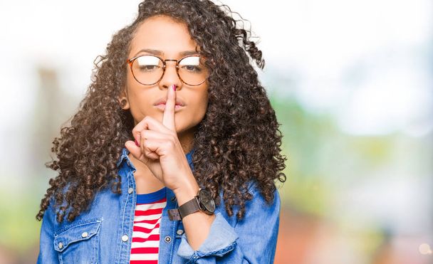 Young beautiful woman with curly hair wearing glasses asking to be quiet with finger on lips. Silence and secret concept. - Photo, Image
