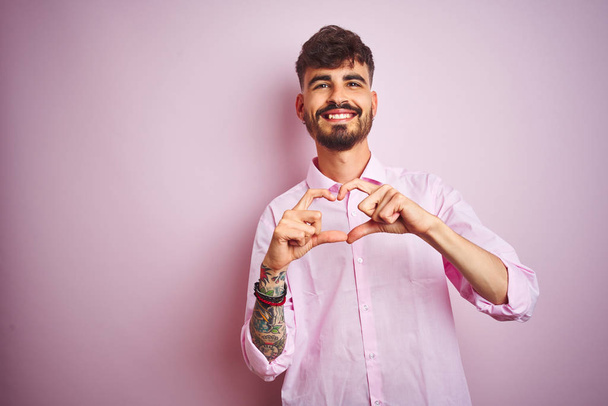 Jovem com tatuagem vestindo camisa em pé sobre fundo rosa isolado sorrindo no amor mostrando símbolo do coração e forma com as mãos. Conceito romântico
. - Foto, Imagem