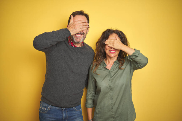 Beautiful middle age couple over isolated yellow background smiling and laughing with hand on face covering eyes for surprise. Blind concept. - Фото, зображення