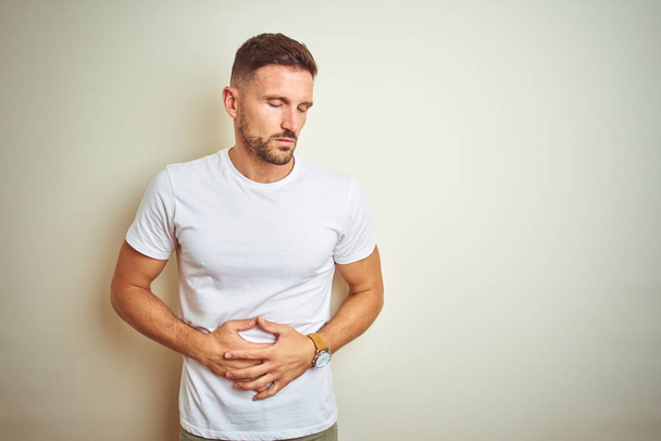 Young handsome man wearing casual white t-shirt over isolated background with hand on stomach because indigestion, painful illness feeling unwell. Ache concept. - Photo, Image
