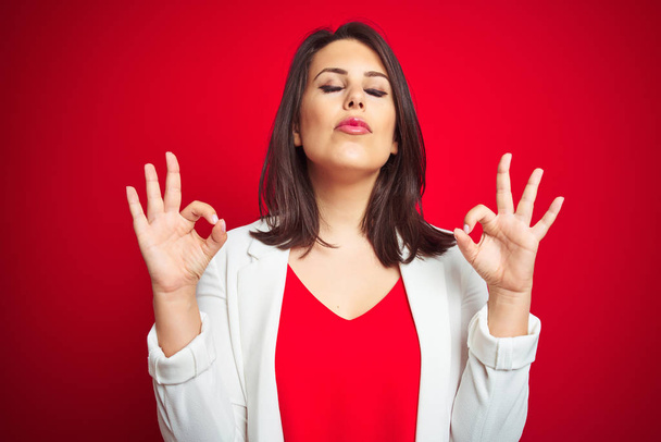 Young beautiful business woman wearing elegant jacket over red isolated background relax and smiling with eyes closed doing meditation gesture with fingers. Yoga concept. - Photo, Image