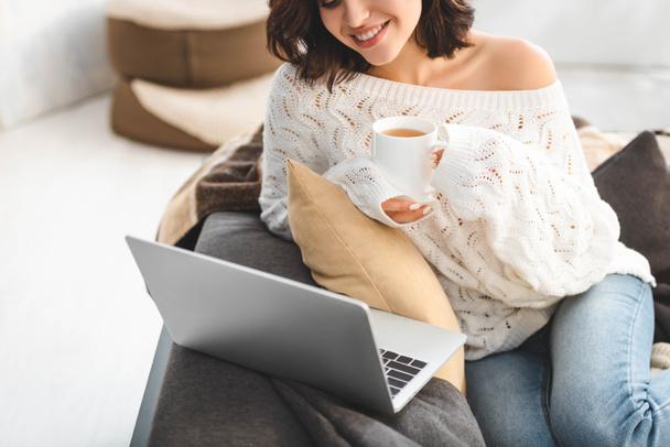 ελκυστική κοπέλα με φλιτζάνι τσάι χρησιμοποιώντας φορητό υπολογιστή στο άνετο σπίτι - Φωτογραφία, εικόνα