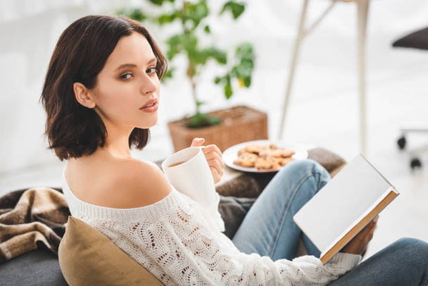 hermosa chica morena leyendo libro en sofá con café y galletas
 - Foto, Imagen