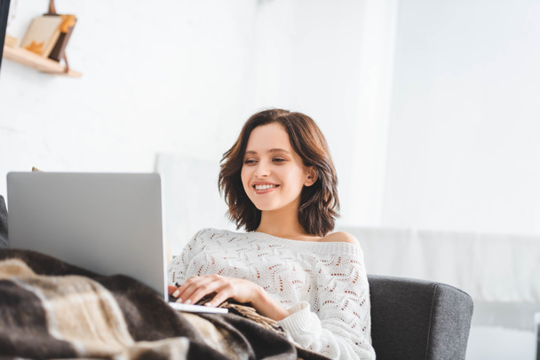 mooie gelukkige vrouw in deken met behulp van laptop op de bank in gezellige woonkamer - Foto, afbeelding