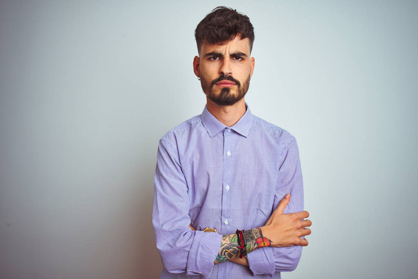 Молодий чоловік з татуюванням у фіолетовій сорочці стоїть над ізольованим білим тлом скептично і нервово, не схвалюючи вираження на обличчі схрещеними руками. Негативна людина
. - Фото, зображення