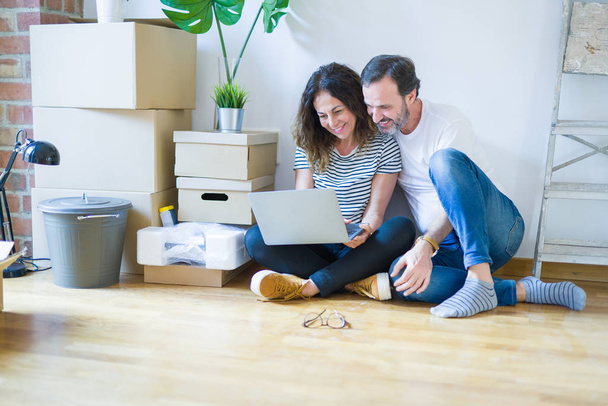 Старшая романтическая пара среднего возраста влюблена, сидя на полу квартиры с коробками вокруг и с помощью компьютера ноутбук улыбается счастливы для переезда в новый дом
 - Фото, изображение
