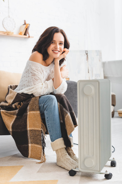 beautiful smiling girl with blanket warming up with heater in cold room - Foto, imagen