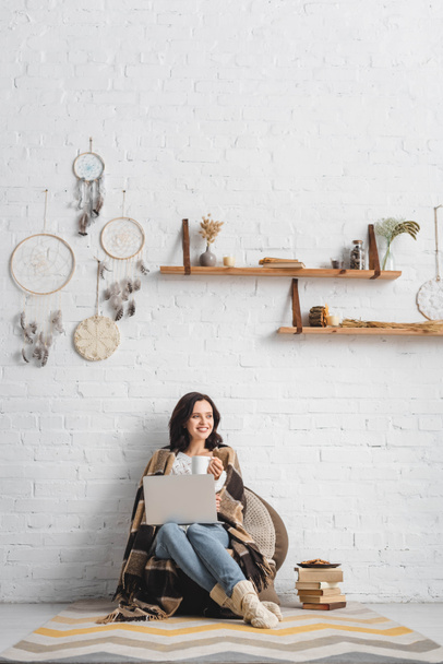 veselá brunetka dívka s cookies a kávou pomocí notebooku v obývacím pokoji s lapači snů - Fotografie, Obrázek
