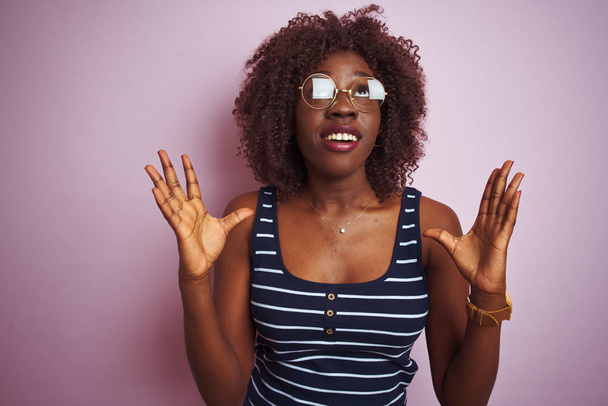 孤立したピンクの背景に縞模様のTシャツの眼鏡をかけている若いアフリカのアフロ女性は、狂気と怒りの叫びと積極的な表現と腕を上げて叫んでいます。欲求不満の概念. - 写真・画像