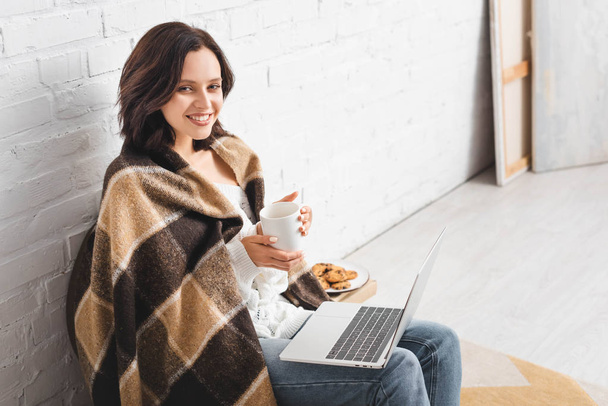 beautiful smiling girl in blanket with cookies and coffee using laptop - Фото, зображення