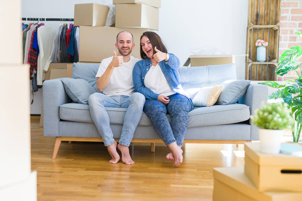 Young couple sitting on the sofa arround cardboard boxes moving to a new house doing happy thumbs up gesture with hand. Approving expression looking at the camera showing success. - 写真・画像