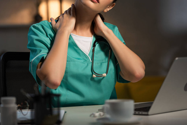 vista recortada de la enfermera en uniforme sentada en la mesa y con dolor de cuello durante el turno de noche
 - Foto, Imagen