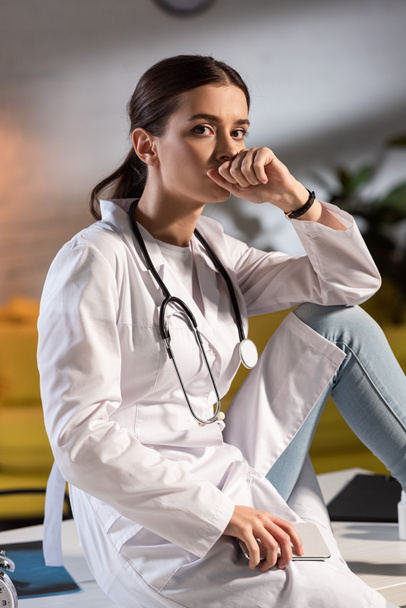 attractive doctor in white coat with stethoscope holding smartphone during night shift - Photo, Image