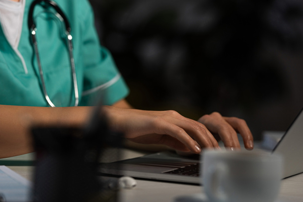 bijgesneden beeld van verpleegster in uniform zitten aan tafel en met behulp van laptop tijdens de nachtdienst - Foto, afbeelding