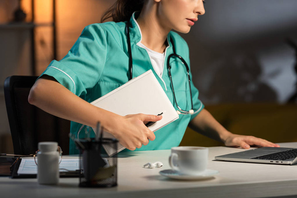 vista recortada de la enfermera en uniforme sentada en la mesa y sosteniendo el cuaderno durante el turno de noche
 - Foto, Imagen