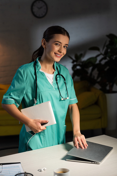 smiling nurse in uniform holding notebook and closing laptop during night shift - Foto, immagini