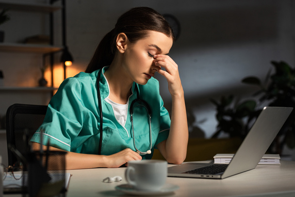 attractive and tired nurse in uniform sitting at table during night shift - Foto, imagen