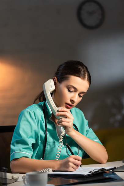 attractive nurse in uniform talking on telephone and writing in clipboard during night shift - Foto, Imagen